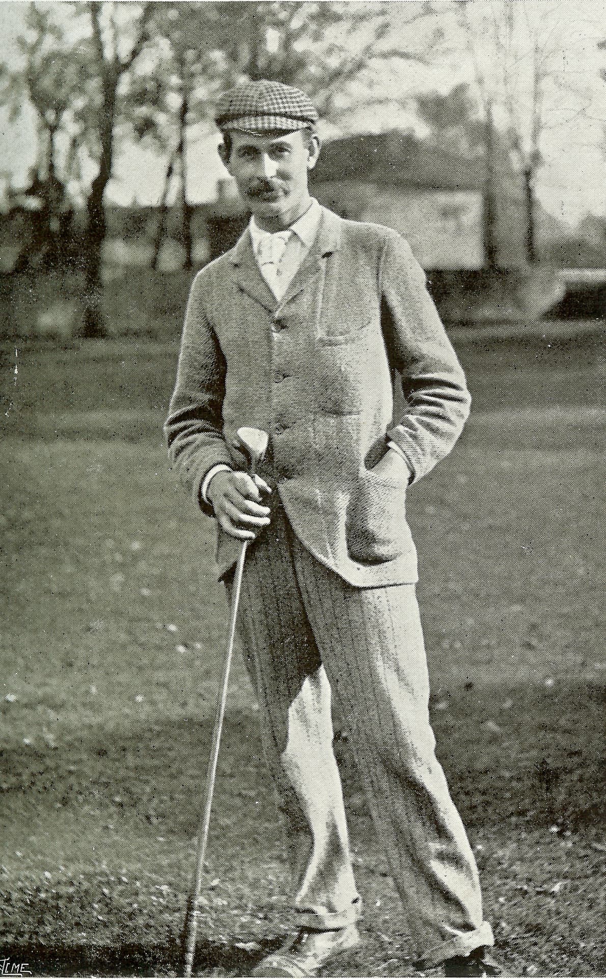Harry Vardon in tweed jacket and cap, standing on turf leaning on a club, 1899