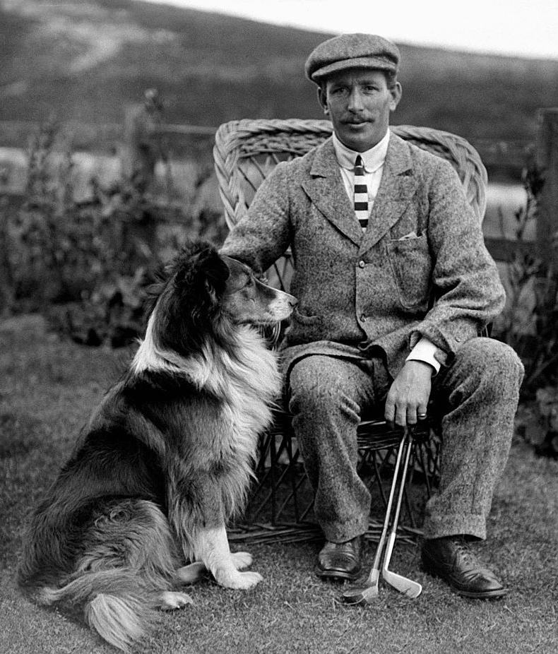 Jack White seated in a wicker chair beside a collie, holding two clubs, 1904