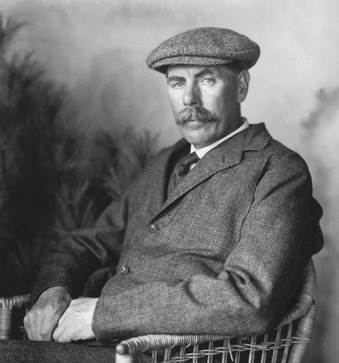 Studio portrait of James Braid seated in a wicker chair wearing a tweed cap and jacket, 1904