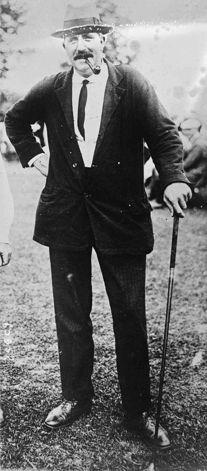 Ted Ray standing with a pipe in his mouth, hand resting on a club, 1920